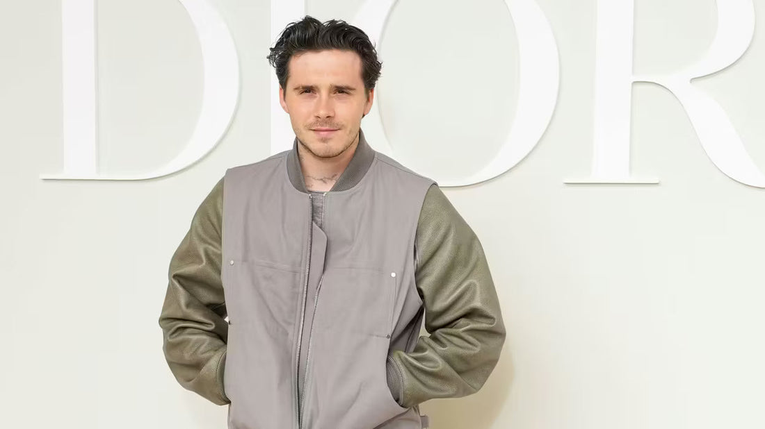 Brooklyn Beckham, wearing a white t-shirt and dark trousers, stands in a modern kitchen preparing a meal on a wooden cutting board.